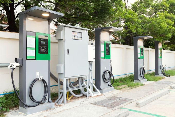 Electric vehicle charging stations with green displays along a sidewalk under trees