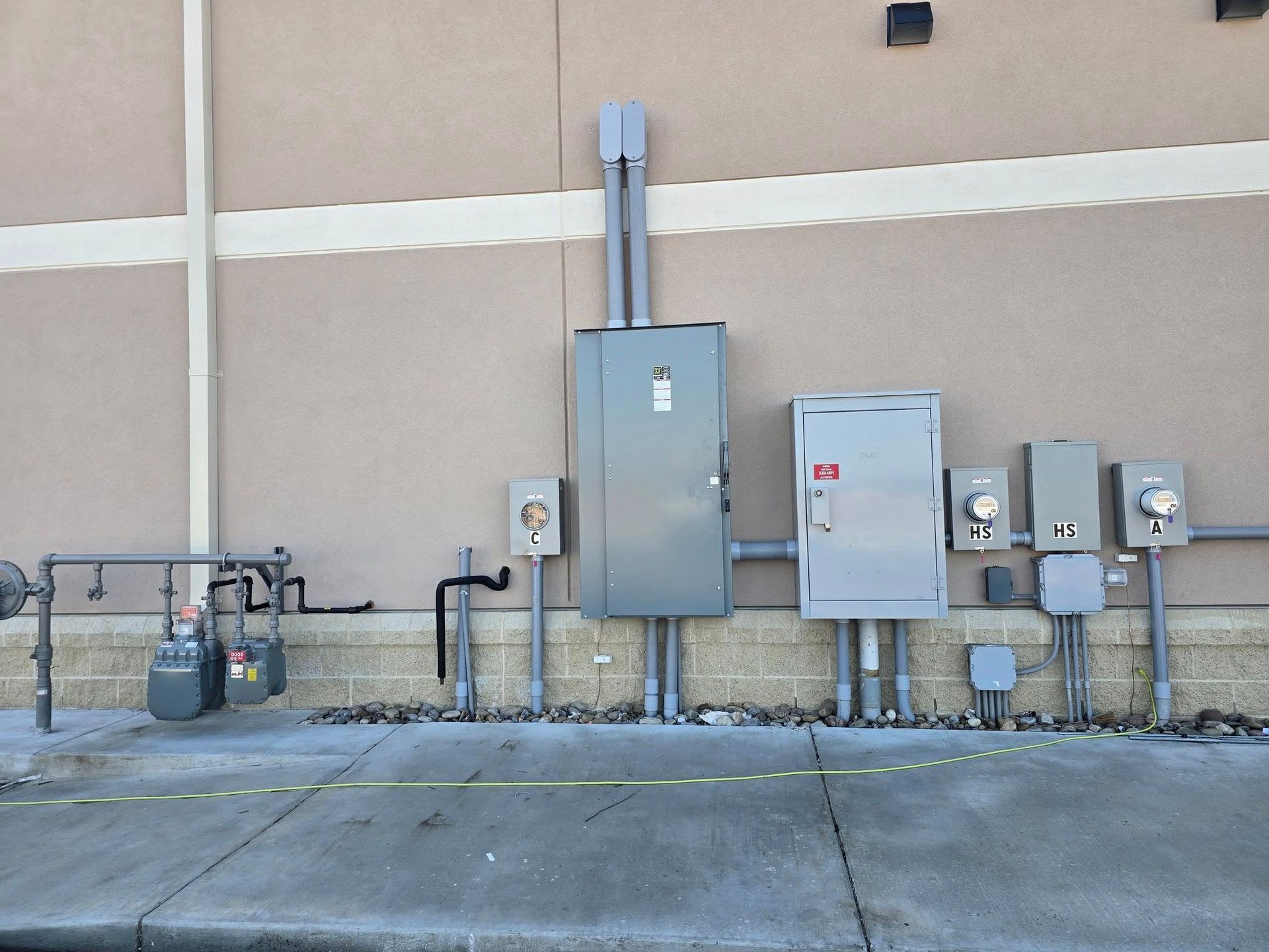 Electrical equipment and gas meter on beige building exterior. Gray metal boxes and pipes. Concrete ground.