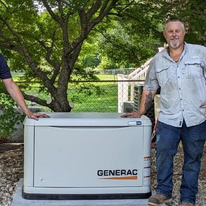 Two men pose beside a Generac generator. One wears jeans and a light shirt, the other is partly visible. Outdoors, by trees.