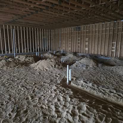 Interior of a building under construction, showing wooden framing, plumbing pipes, and sandy ground.