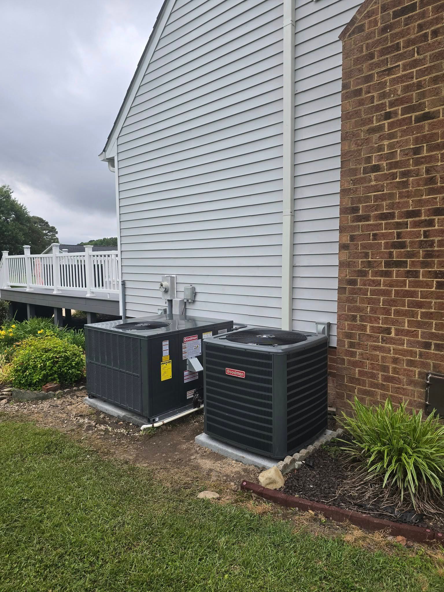 Two air conditioning units beside a white-sided house and a brick chimney, sitting on concrete pads. Green grass surrounds.