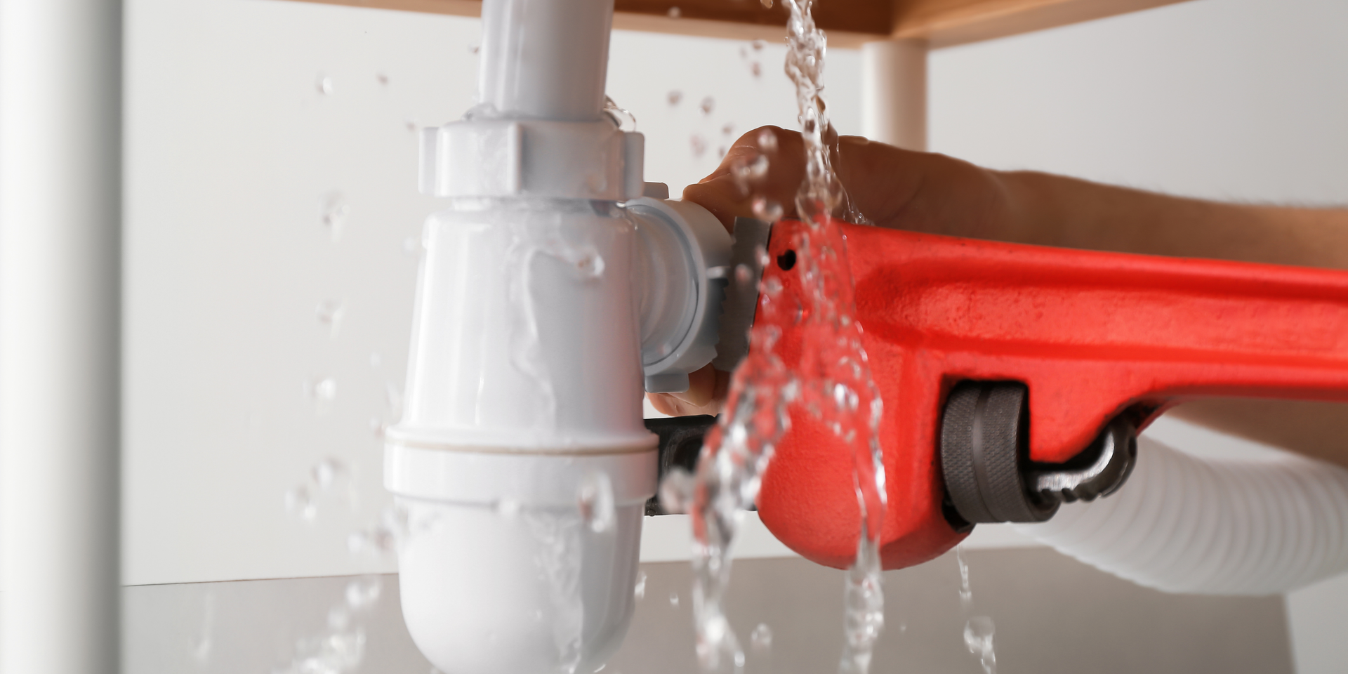 A red wrench is used on a leaking sink pipe. Water streams out as the pipe is worked on.