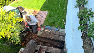 Worker using a jackhammer to break concrete beside a lawn and house exterior