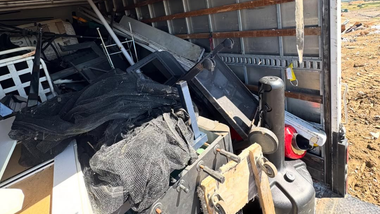 Damaged black furniture and debris piled inside a cluttered garage, with a red bucket and tools nearby