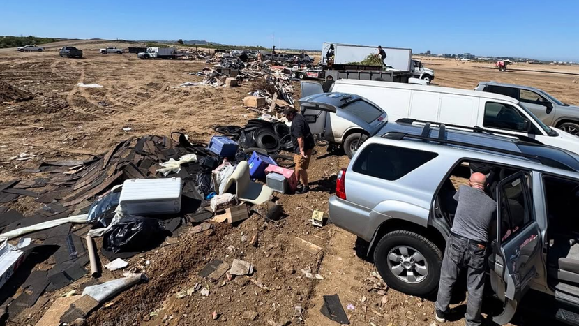 Damaged vehicles and debris scattered across a dry disaster area with people inspecting wreckage