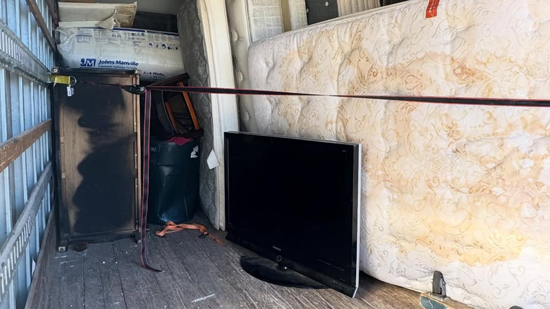 A cluttered storage area with a large black TV leaning against a wall beside stacked equipment.