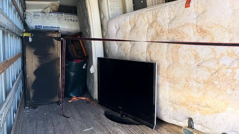 A cluttered storage area with a large black TV leaning against a wall beside stacked equipment.