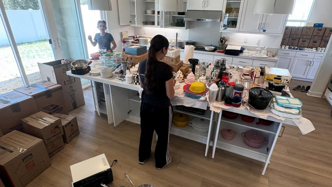 Two people organizing supplies on a kitchen island in a bright kitchen, with boxes stacked nearby.