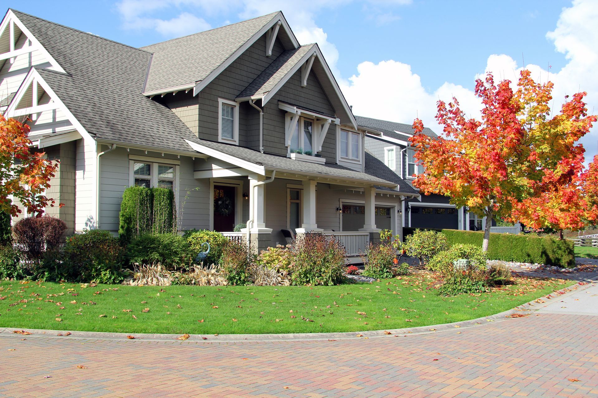 House with gray roof, white trim, porch, and vibrant autumn trees. Green grass, brick driveway.