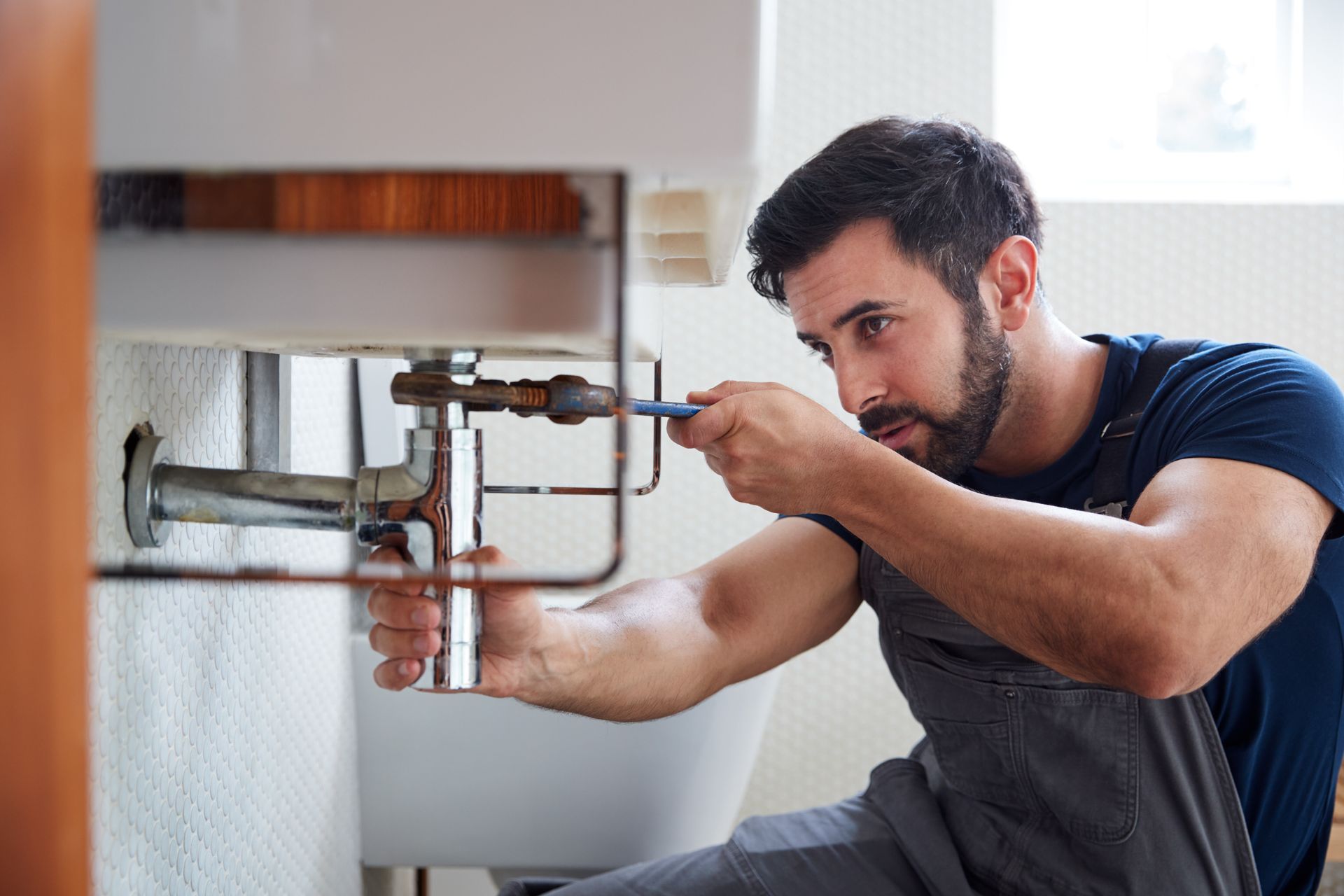 Male plumber wearing a blue uniform and gloves, using a wrench to repair a leaking sink in a home bathroom.