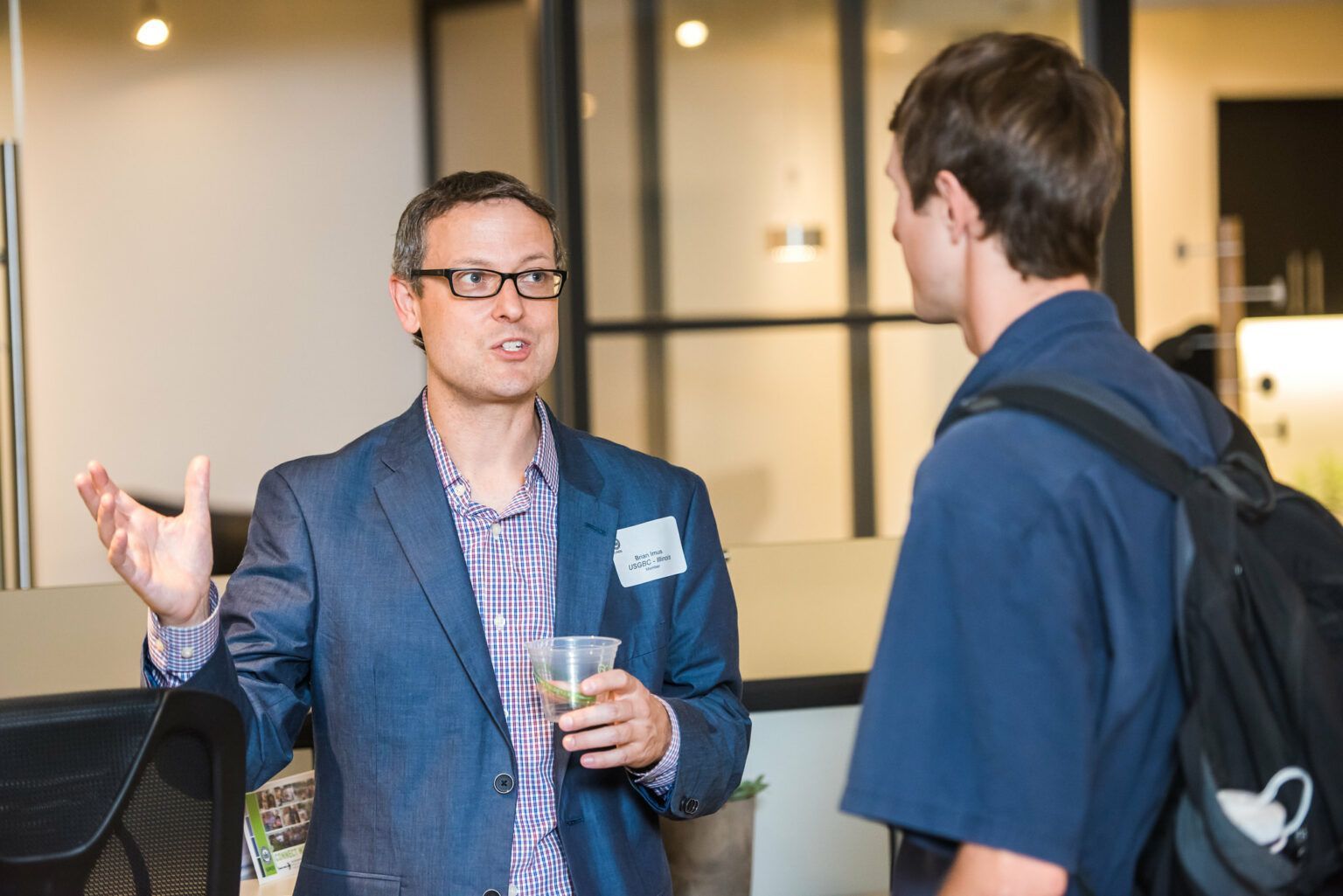 A man in a suit and glasses is talking to another man in a blue shirt.