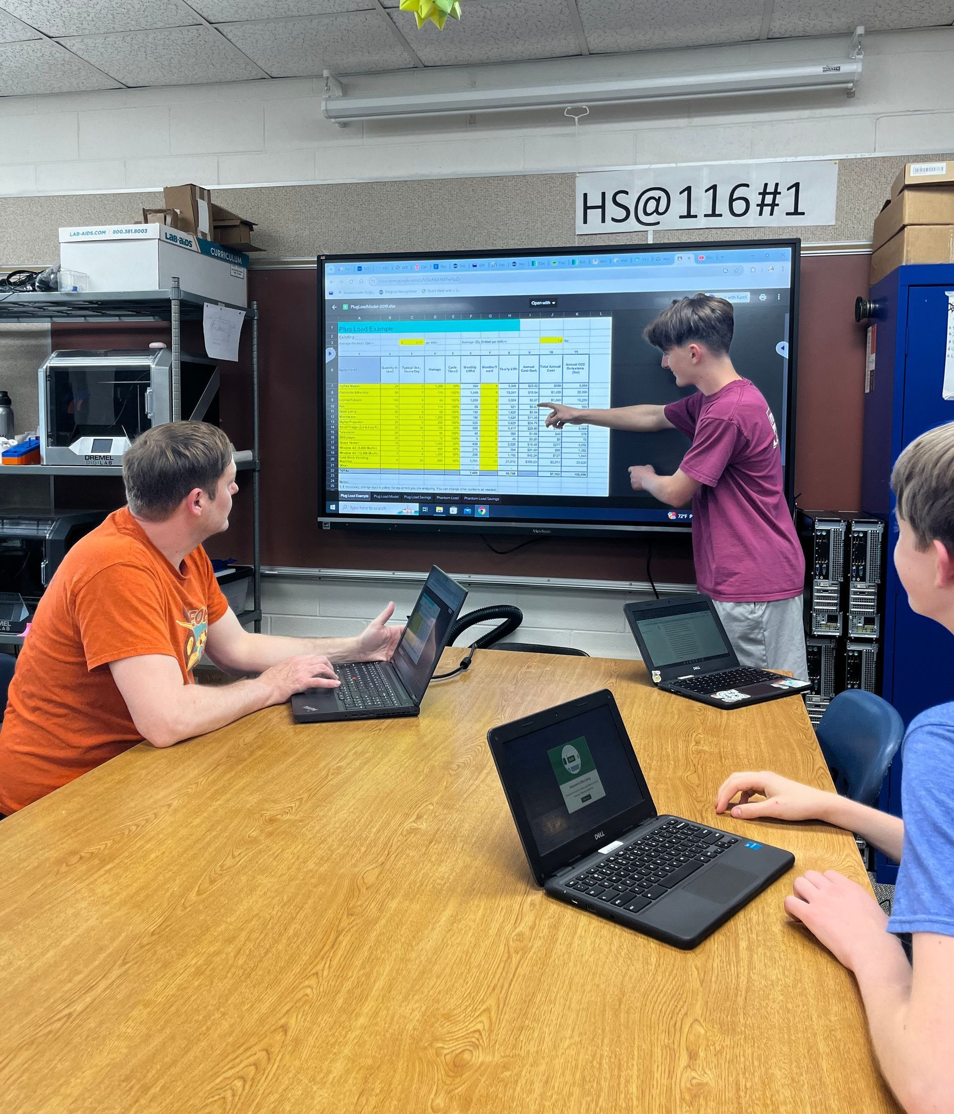 A group of people are sitting at a table with laptops in front of a large screen.