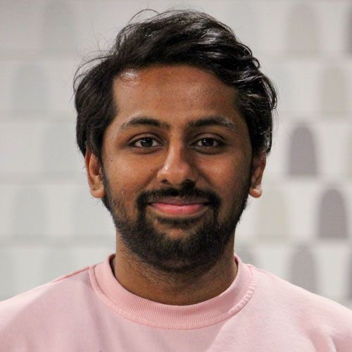 A man with a beard is wearing a pink sweater and smiling.