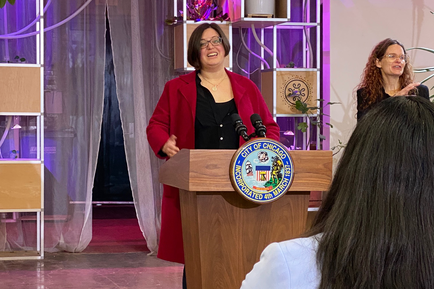 A woman in a red coat is standing at a podium giving a speech.