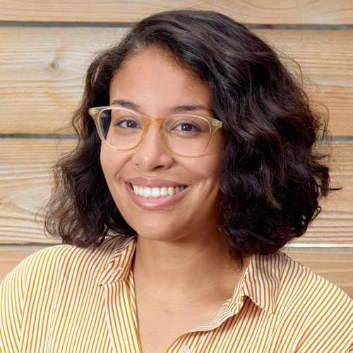 A woman wearing glasses and a striped shirt is smiling in front of a wooden wall.