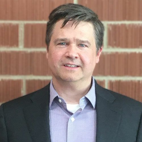 A man in a suit and plaid shirt is standing in front of a brick wall.