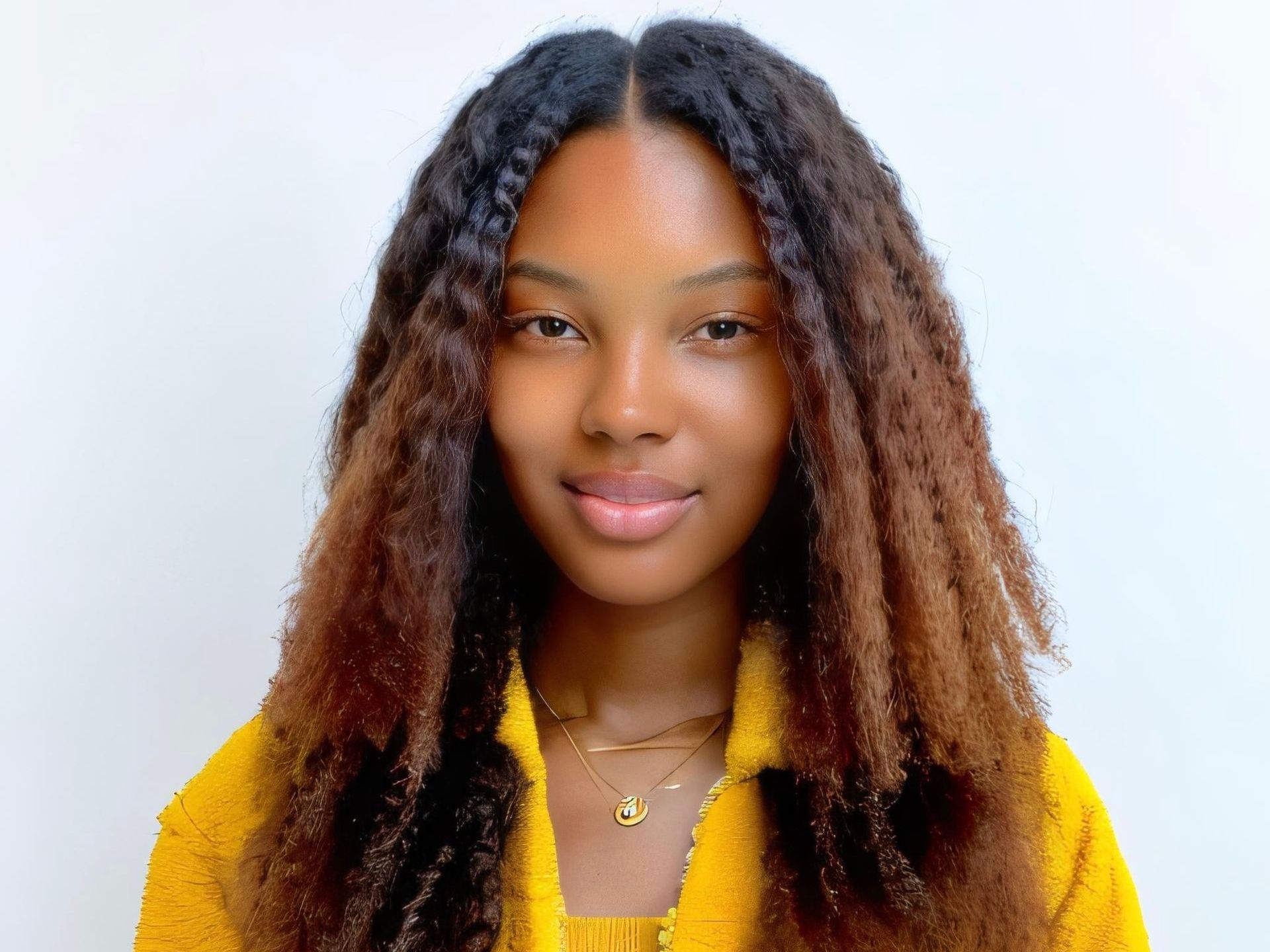 A woman with long curly hair is wearing a yellow jacket and a necklace.