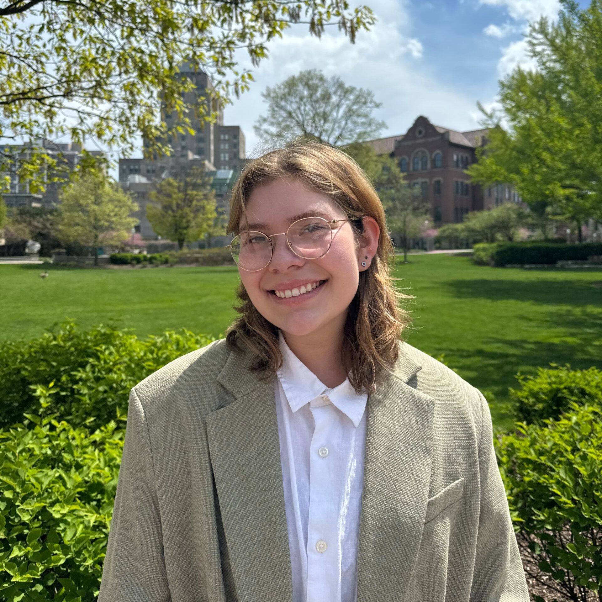 A young woman wearing glasses and a jacket is standing in a park.