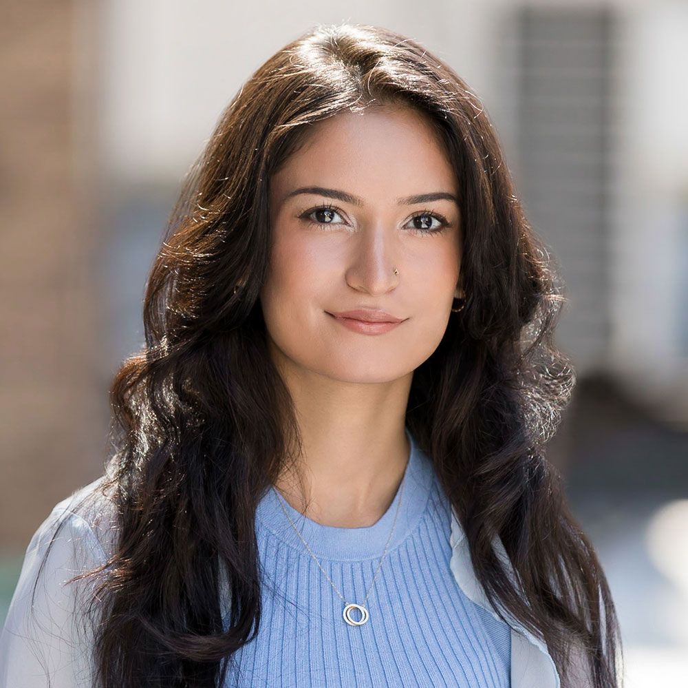 A woman with long dark hair is wearing a blue sweater and a grey jacket.