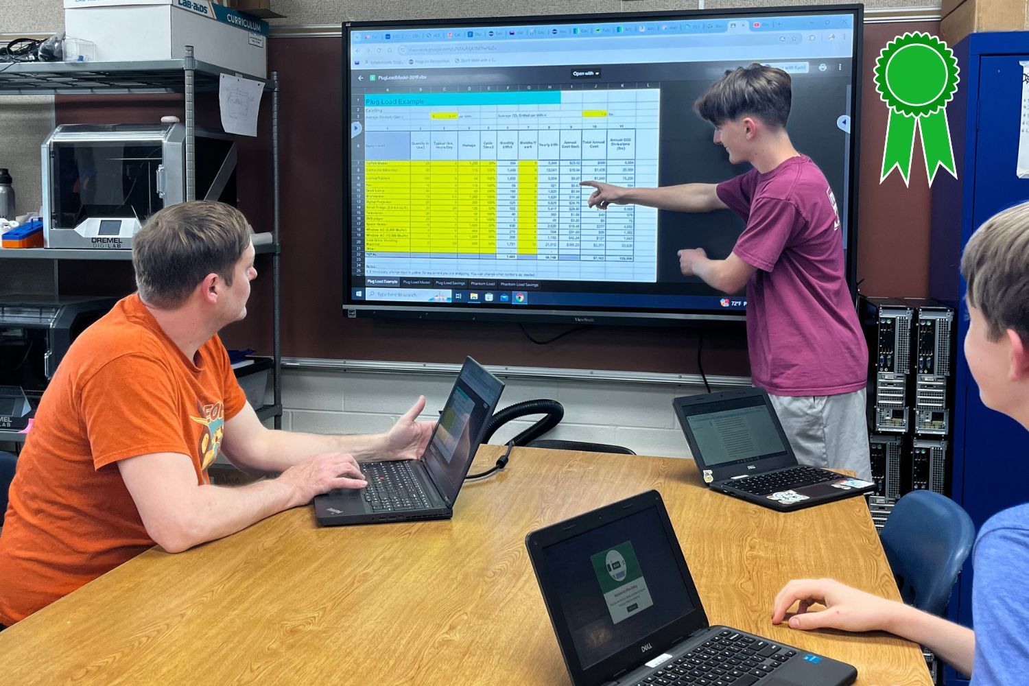A group of people are sitting at a table with laptops in front of a large screen.