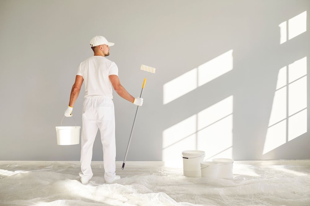 Painter in white overalls stands in a room, holding a roller and a bucket, preparing to paint a wall.