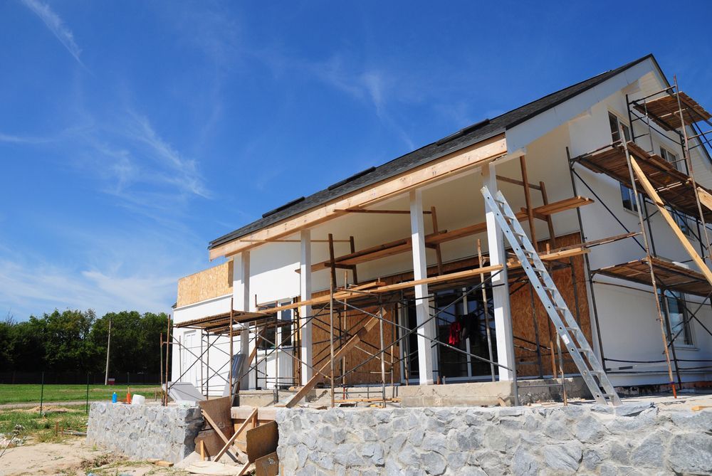 House under construction with scaffolding, blue sky, white walls, stone foundation.