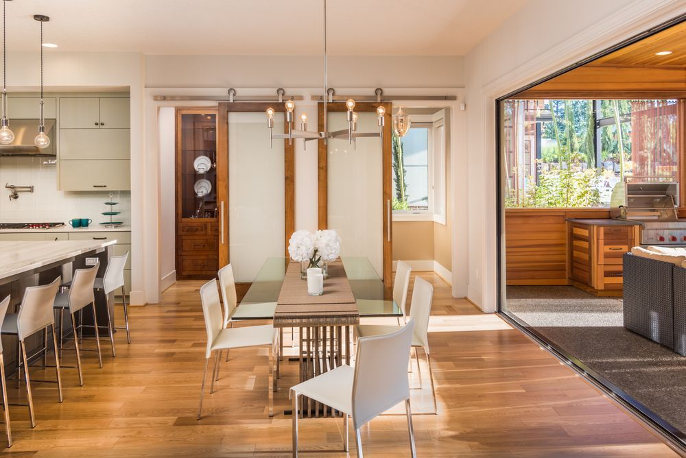 Bright dining room with glass table, white chairs, open to a kitchen and outdoor space.