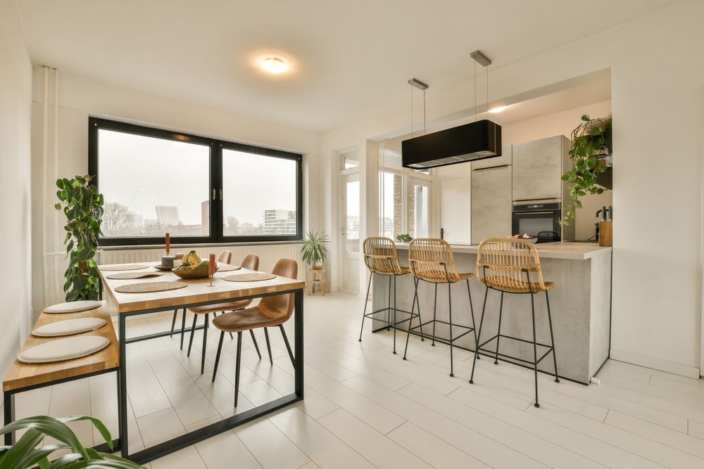 Bright dining area with table, bench, chairs, and kitchen island with stools; plants throughout.