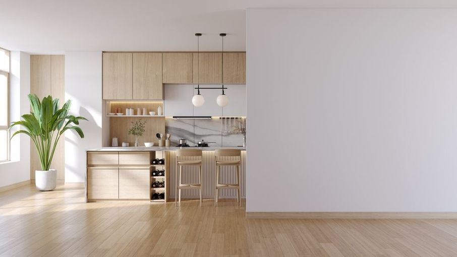 Modern kitchen interior with wooden cabinets, island, and stools. A large plant and a white wall are also visible.