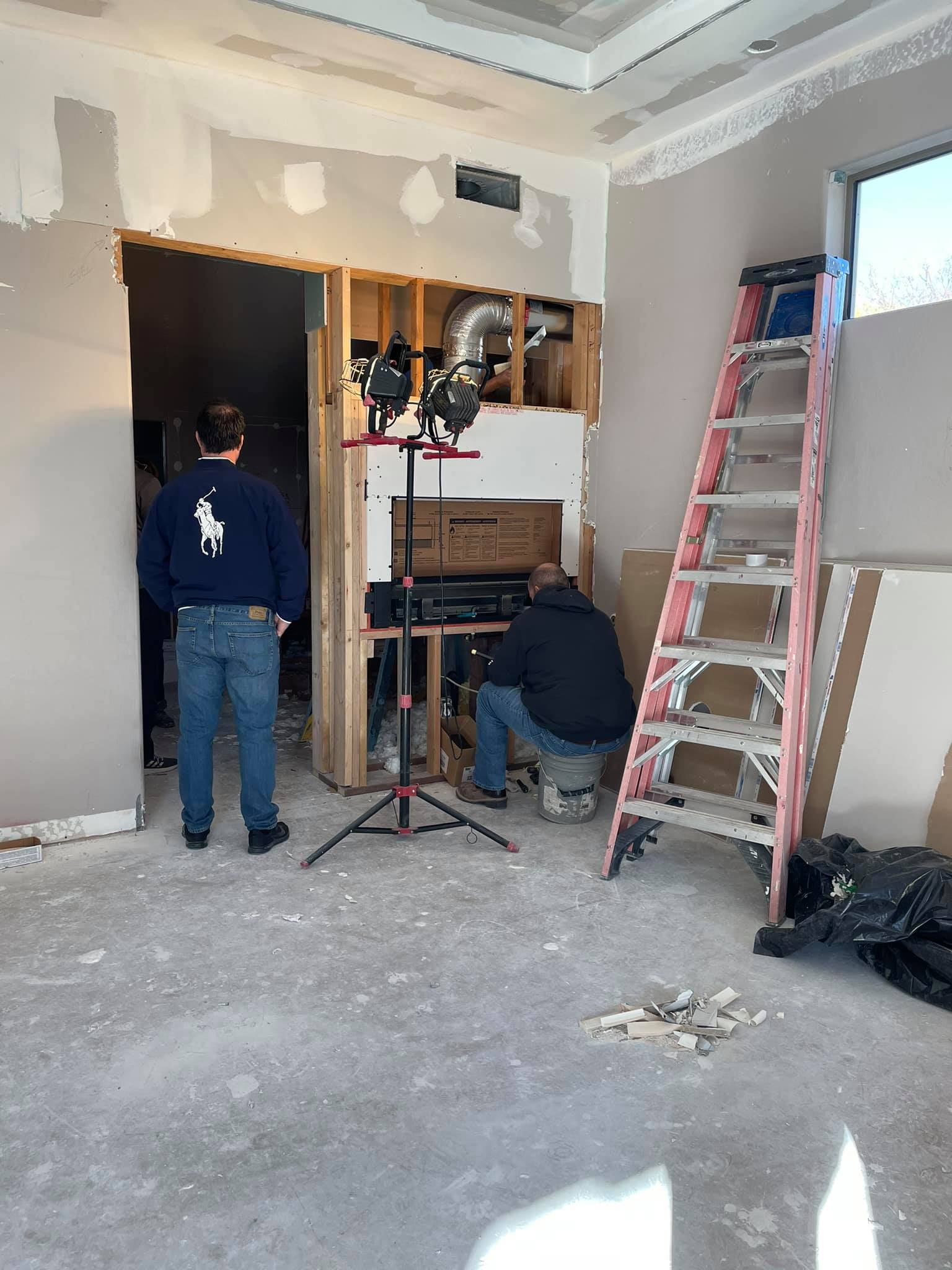 Two men working on a room remodel near a fireplace. One stands, one kneels. Pink ladder, exposed framing.