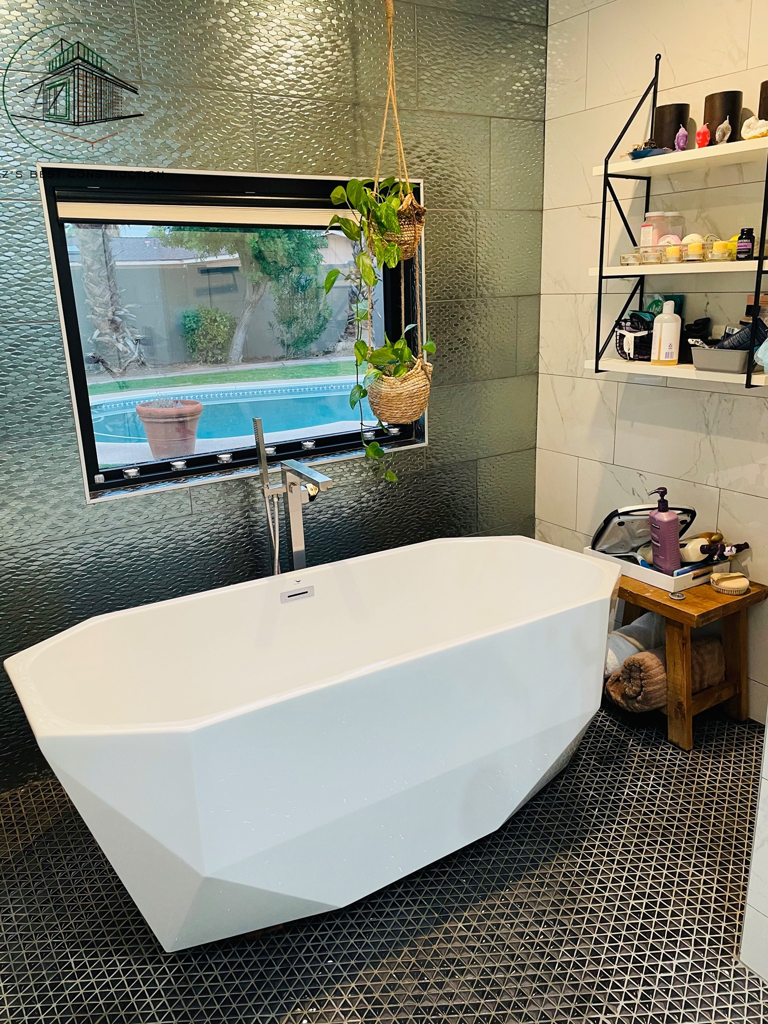 White geometric bathtub in a modern bathroom with textured green tile wall and a window overlooking a pool.