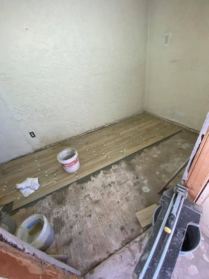 A room with flooring being installed. Wood planks are partially laid on concrete floor. Buckets, tool.