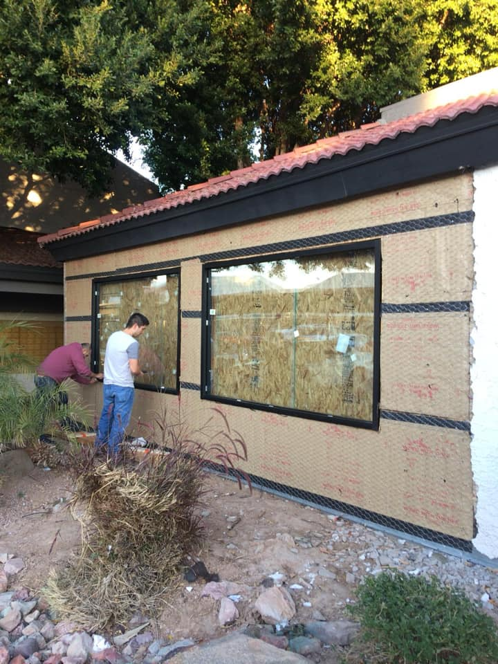 Two people working on a house exterior. Brown siding, black window frames. Green trees and bushes in front.