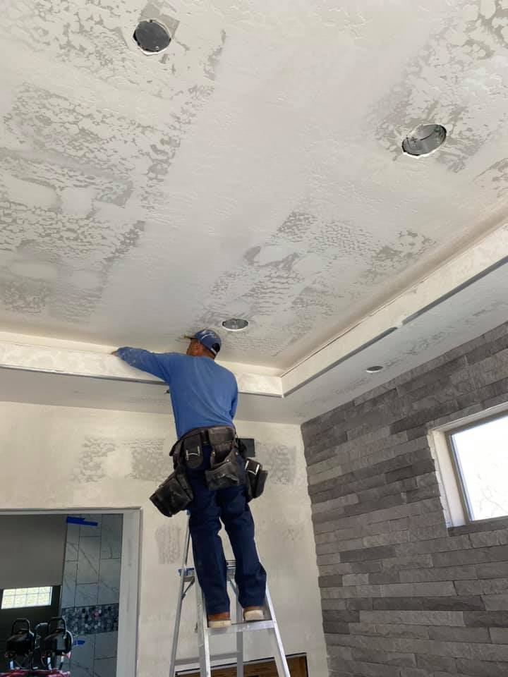 Man on a ladder installing trim on a white ceiling with recessed lights, gray stone wall.