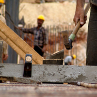 Construction worker hammering wood on a building site, other workers visible in the background.