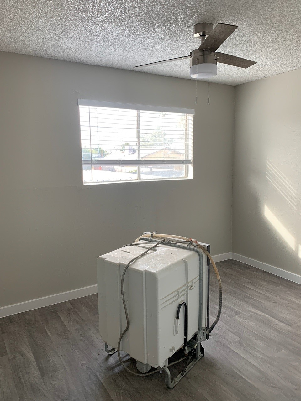 White appliance on wheels in a room with gray walls, a window, and gray wood-look flooring.
