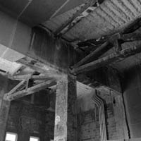 Interior view of a decaying building with wooden beams, columns, and exposed electrical conduits. Black and white.