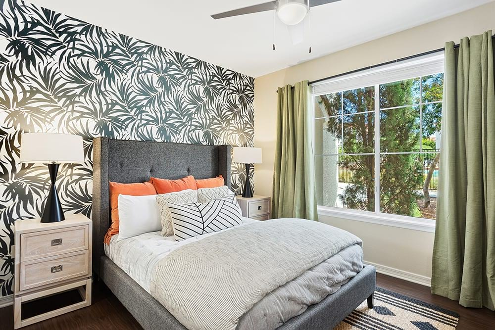 Bedroom with patterned accent wall, gray bed, lamps, window with green curtains.