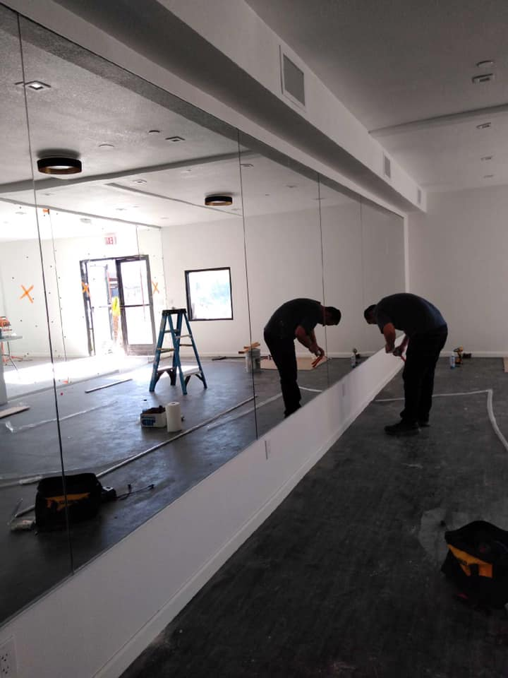 Two workers installing a long white trim piece on a wall with a large mirror.