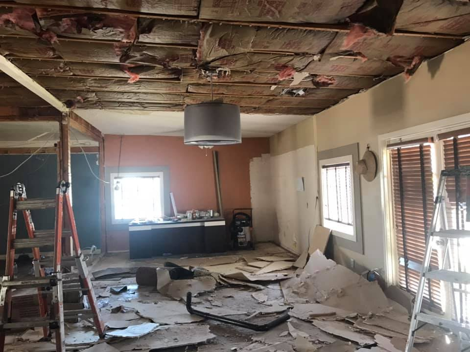 Interior during renovation: damaged ceiling, debris on floor, exposed wood, ladders, walls, light fixture.