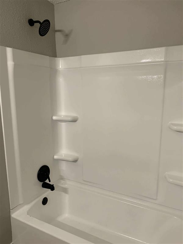 White bathtub and shower with black fixtures, two shelves, and gray wall.