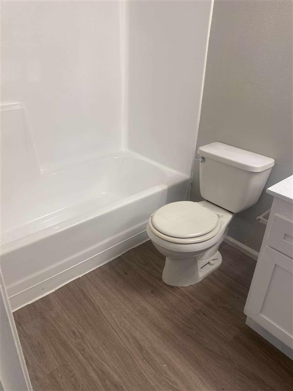 Bathroom with white tub/shower, toilet, white vanity, and wood-look flooring. Gray wall.