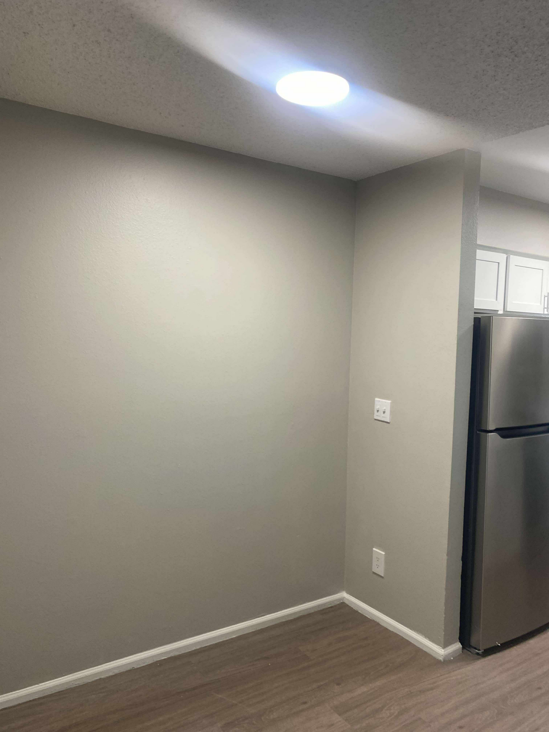 Empty corner of a room with grey walls, white trim, and a stainless steel refrigerator visible.