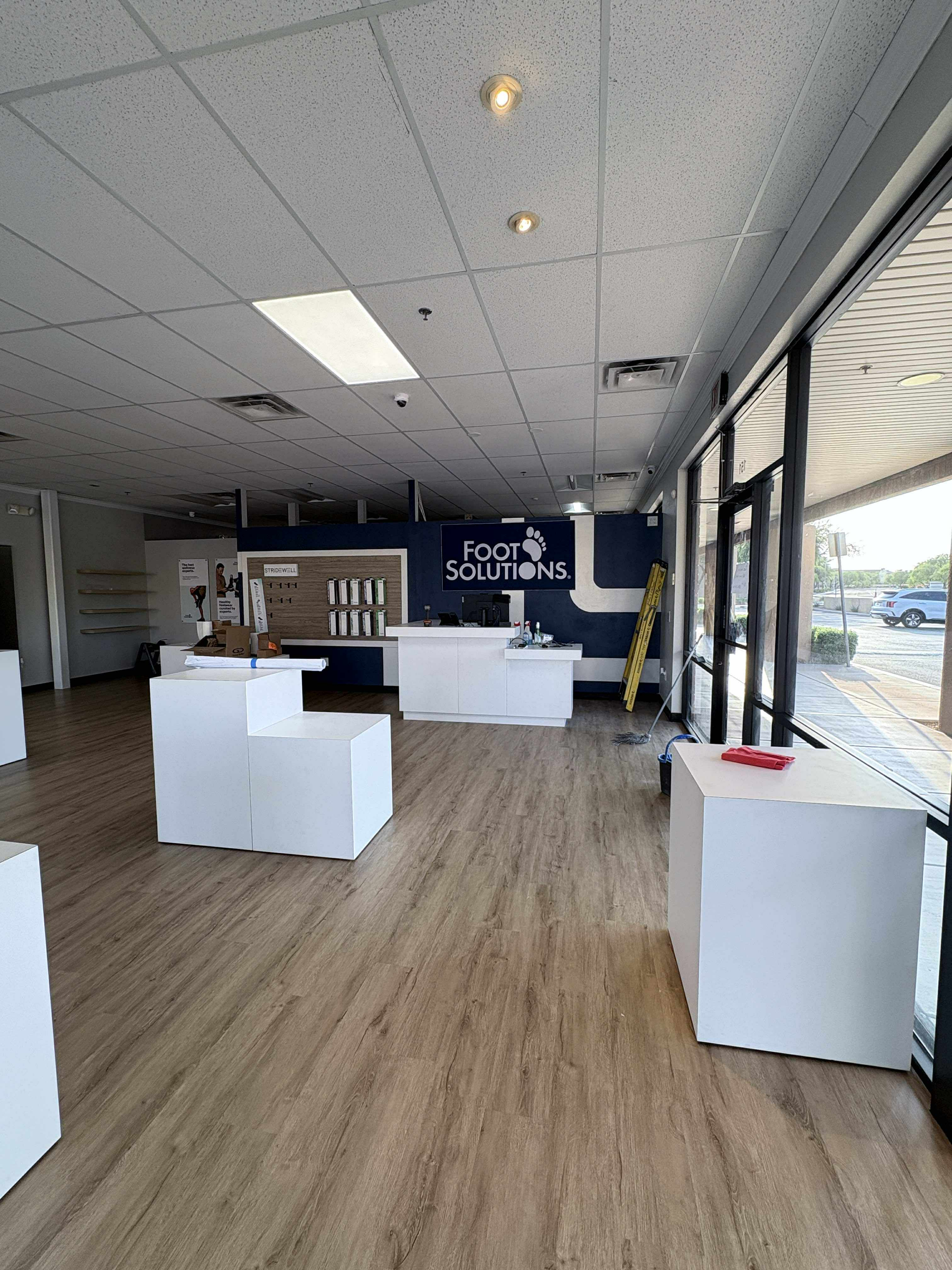 Empty retail space with white displays and fixtures. Light-colored wood flooring, large windows, and a sign for 