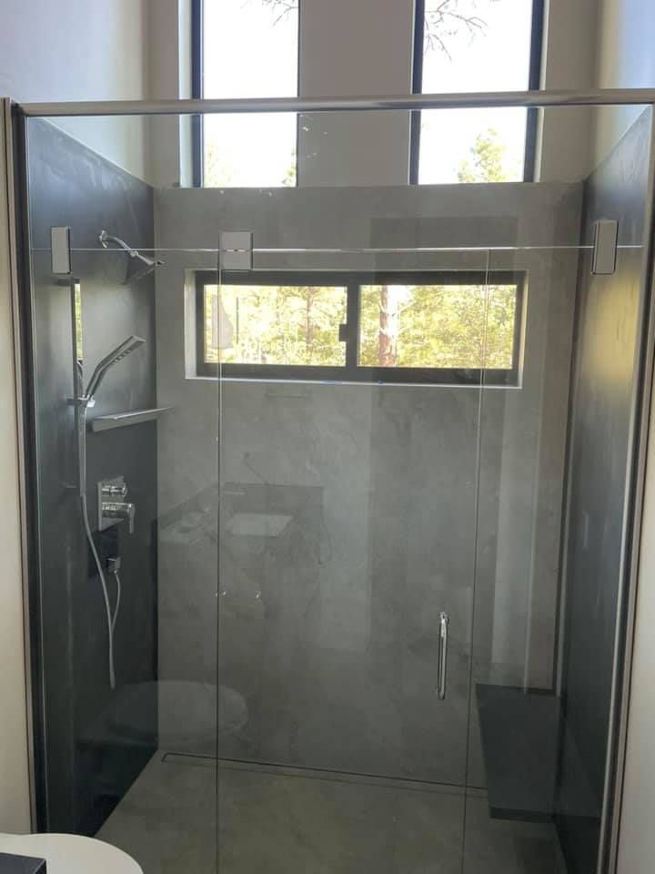 Modern shower with glass walls and door, gray tile, silver fixtures, and a small window overlooking trees.
