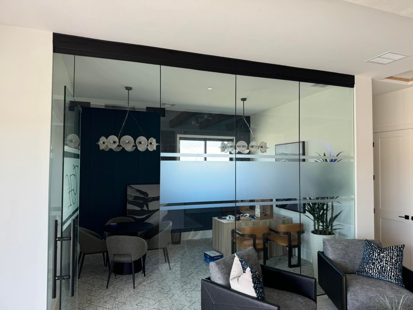 Glass-walled office with frosted horizontal band; black frame; chairs, table, and plant visible inside.