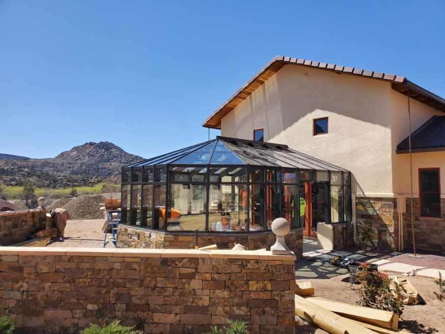Sunroom attached to a beige house, with glass walls and black framing. Brown stone wall and mountain backdrop.