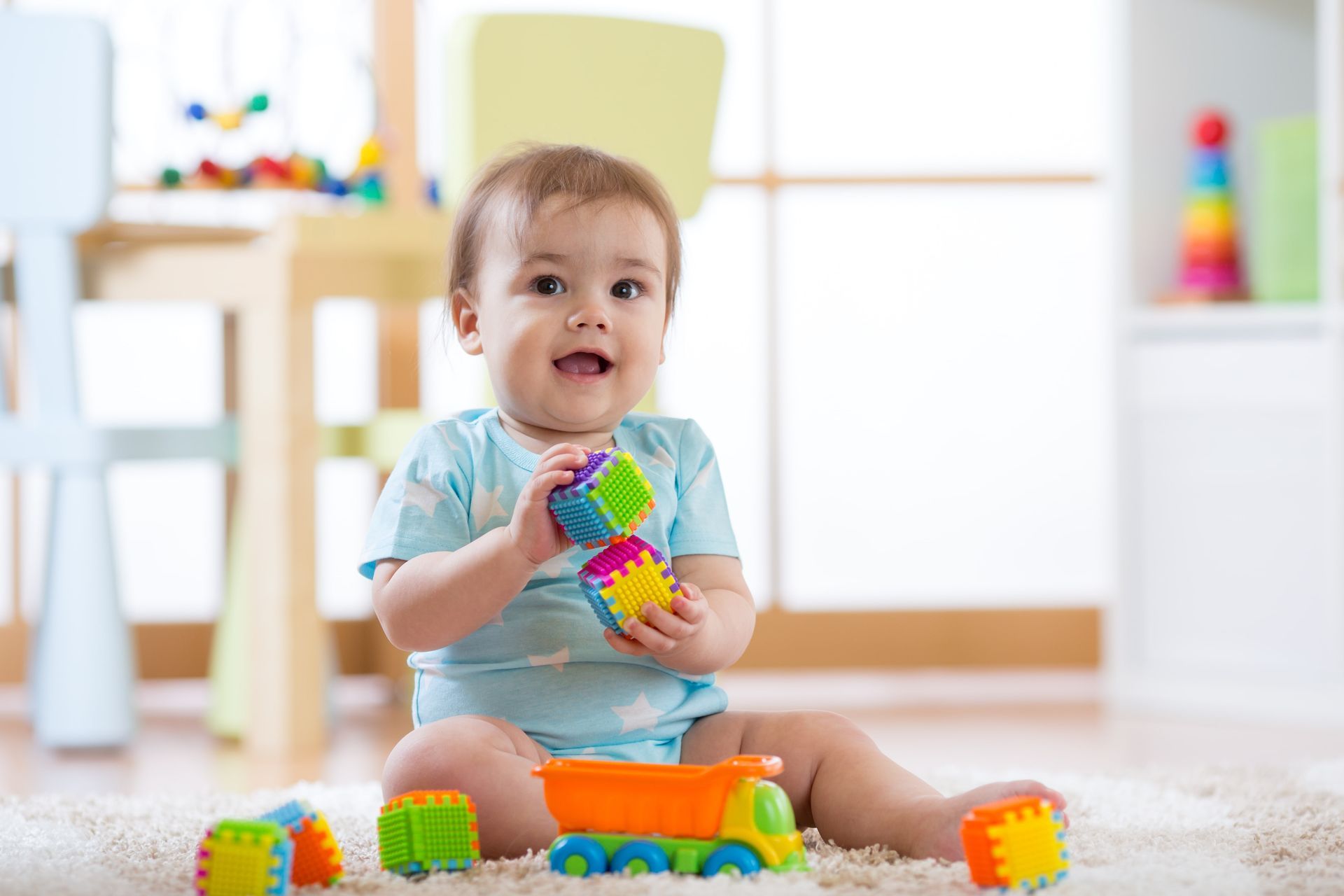 Baby sitting on a rug playing with colorful stacking blocks and a toy truck in a bright room.
