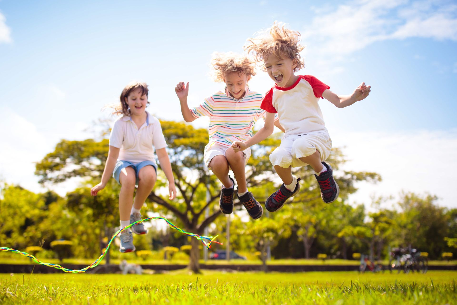 Three children jumping rope on green grass in sunlight.
