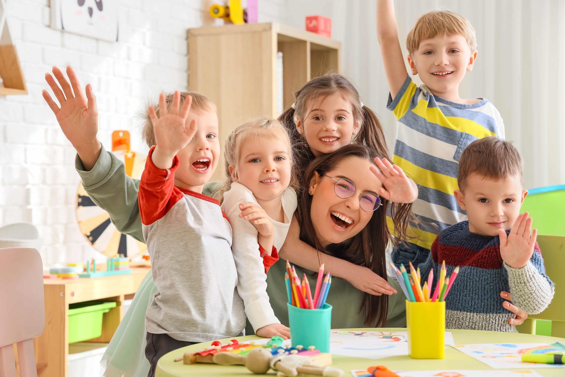 Children and teacher smiling and waving at a table with crayons in a brightly lit classroom.